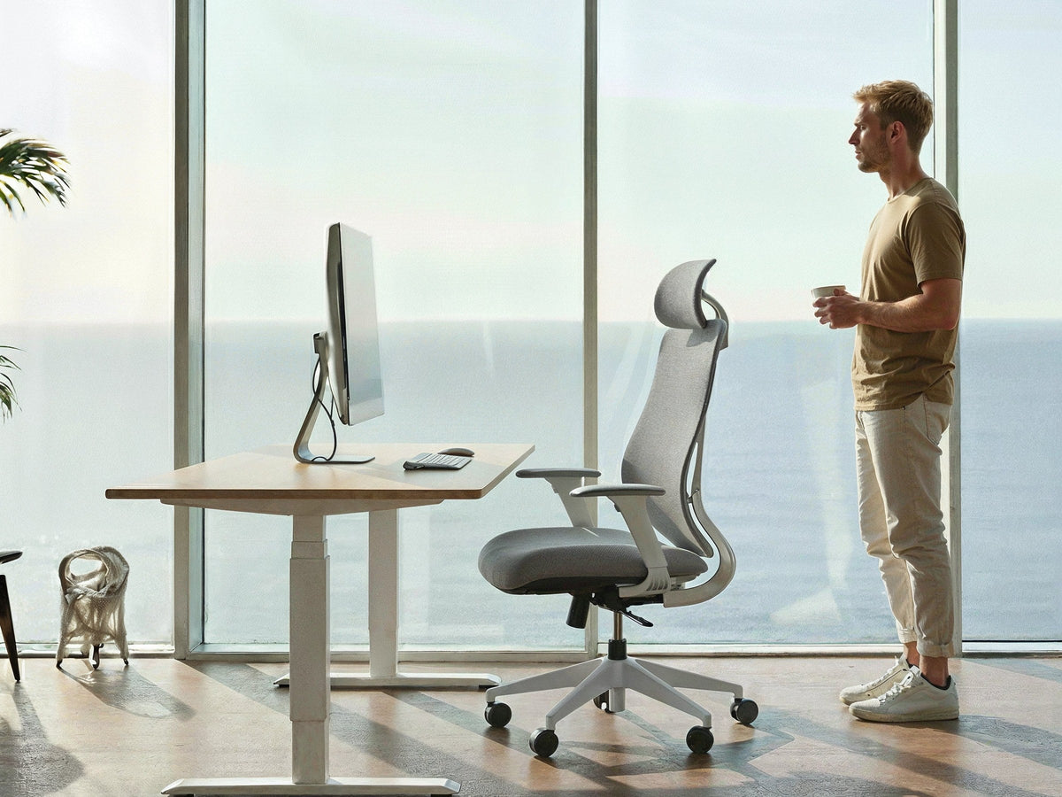 Ergonix regulējama augstuma galds un OmniSit Pro krēsls pie liela loga, blakus stāv vīrietis ar krūzi