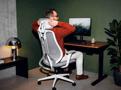 Vīrietis, sēžot Ergonix OmniSit Pro krēslā, regulē galvas balstu, skats no mugurpuses