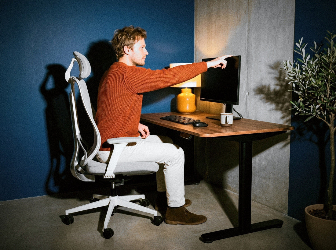 Vīrietis, sēžot Ergonix OmniSit Pro krēslā, ar izstieptu roku sniedzas līdz monitoram: attālums līdz ekrānam – rokas garumā