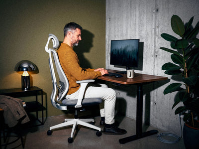 Vīrietis sēž Ergonix OmniSit Pro krēslā pie regulējama augstuma galda: pēdas uz grīdas, elkoņi galda līmenī, ekrāna augšmala acu augstumā